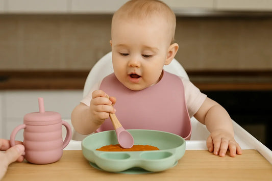 Bébé utilise une cuillère en bois et silicone pour manger une purée dans une assiette compartimentée, avec bavoir et tasse assortis.