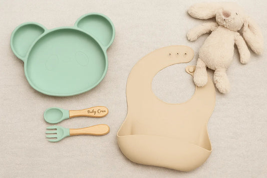 Set repas bébé pastel avec assiette panda en silicone, couverts bois et silicone gravés, bavoir beige et doudou lapin sur fond naturel.