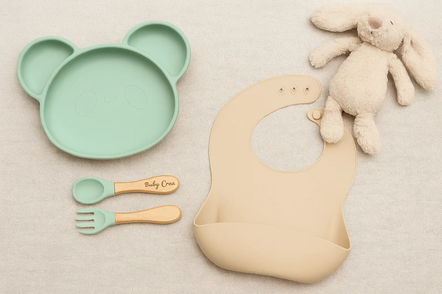 Set repas bébé pastel avec assiette panda en silicone, couverts bois et silicone gravés, bavoir beige et doudou lapin sur fond naturel.