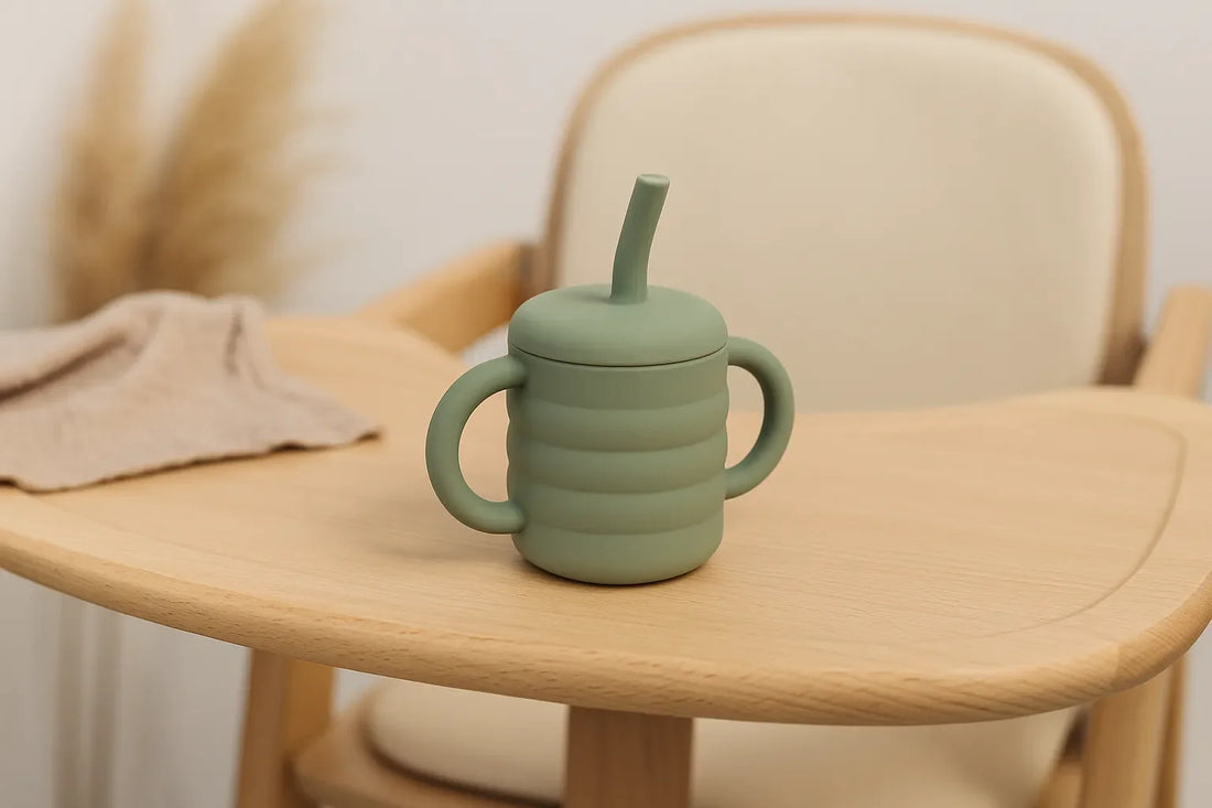 Tasse d’apprentissage bébé en silicone vert avec poignées et paille, posée sur une chaise haute en bois dans un décor naturel.
