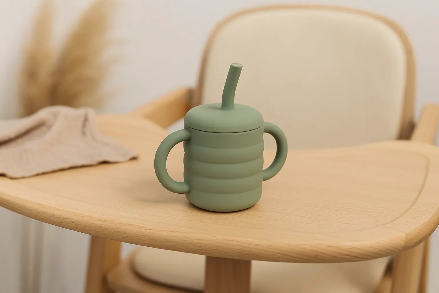 Tasse d’apprentissage bébé en silicone vert avec poignées et paille, posée sur une chaise haute en bois dans un décor naturel.