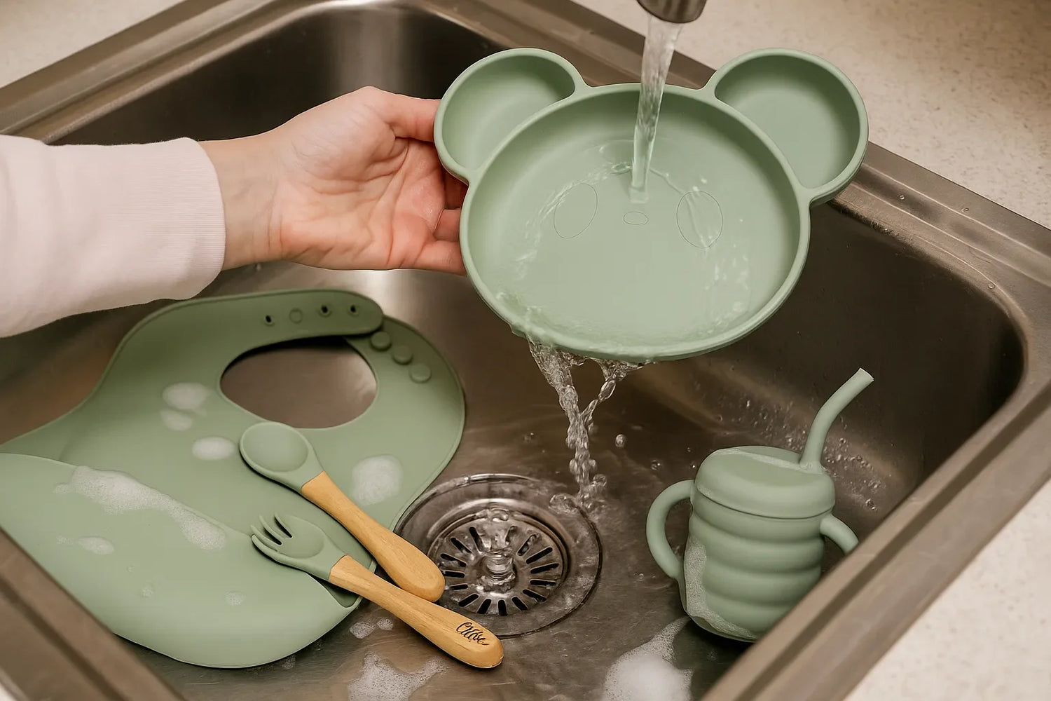 Vaisselle bébé en silicone vert lavée à la main : assiette panda, bavoir, tasse et couverts dans un évier inox avec mousse et eau claire.