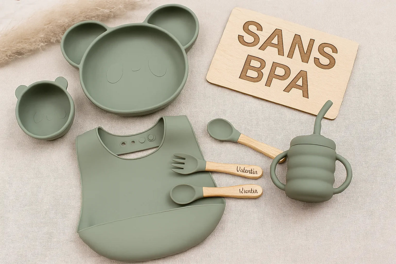 Vaisselle bébé sans BPA : assiette panda, bol, tasse, couverts gravés et bavoir en silicone vert sur fond beige avec panneau “SANS BPA”.