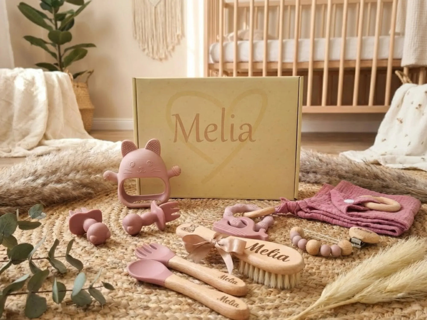 Coffret de naissance décoré en rose, affichant divers accessoires dont une brosse, des couverts et un jouet, entouré d'un décor chaleureux, avec le nom Mélia.