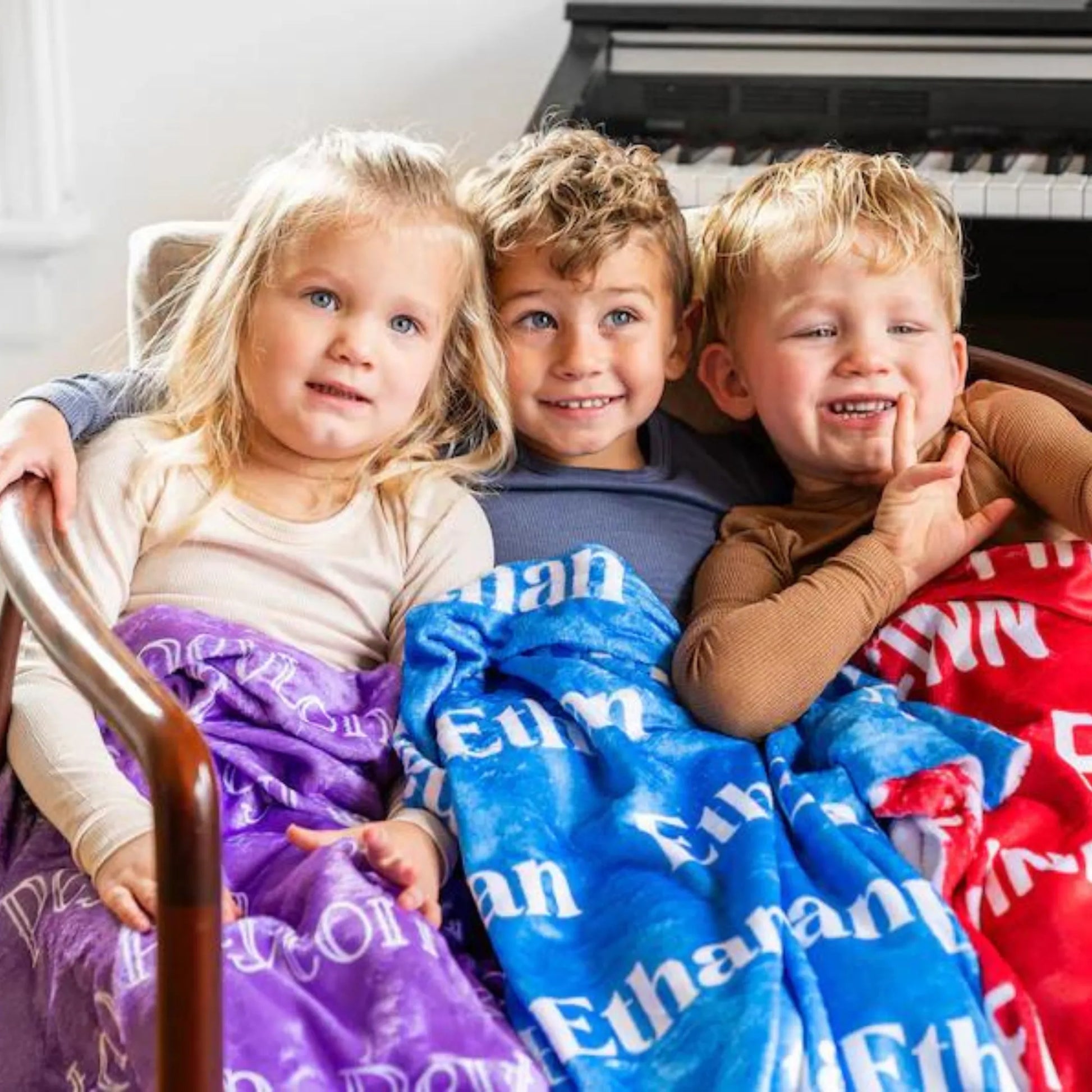 Trois jeunes enfants assis ensemble sur un fauteuil, chacun couvert par son propre plaid personnalisé en violet, bleu roi et rouge vif.