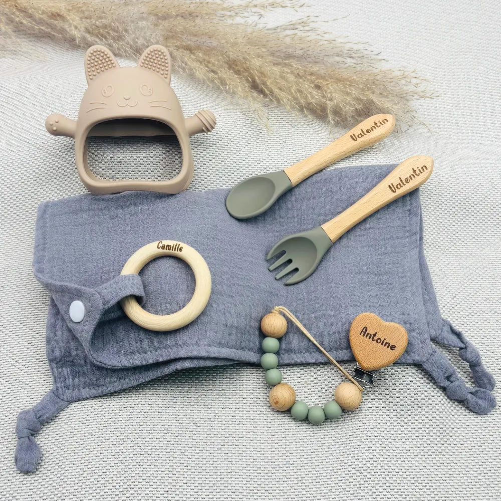 Set repas bébé personnalisé avec bavoir lange gris, couverts en bois gravés Valentin, attache tétine prénom Antoine, anneau de dentition en bois prénom Camille, jouet de dentition chat en silicone beige – cadeau de naissance original et pratique.