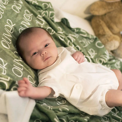 Bébé éveillé allongé sur un plaid de naissance personnalisé vert olive prénom Oliver. Tissu minky doux et résistant pour le confort de l'enfant.