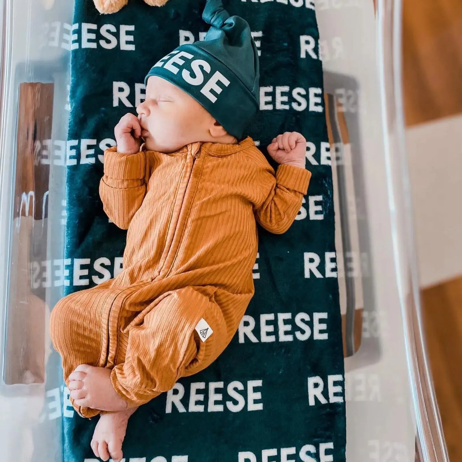 Ensemble cadeau naissance : couverture personnalisée verte prénom Reese avec bonnet assorti. Bébé en pyjama moutarde dormant paisiblement à la maternité.